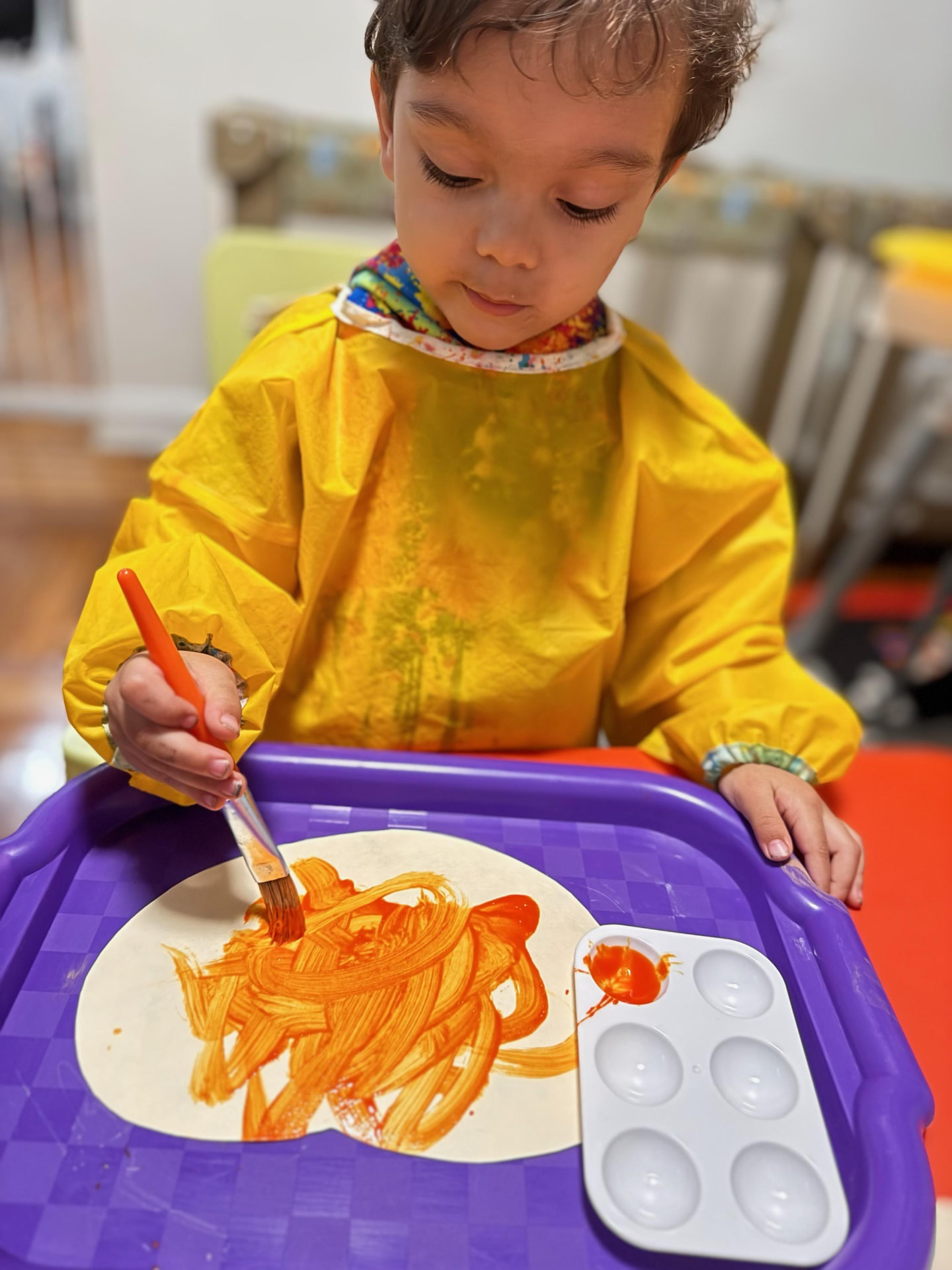 A toddler in a yellow smock painting with orange paint