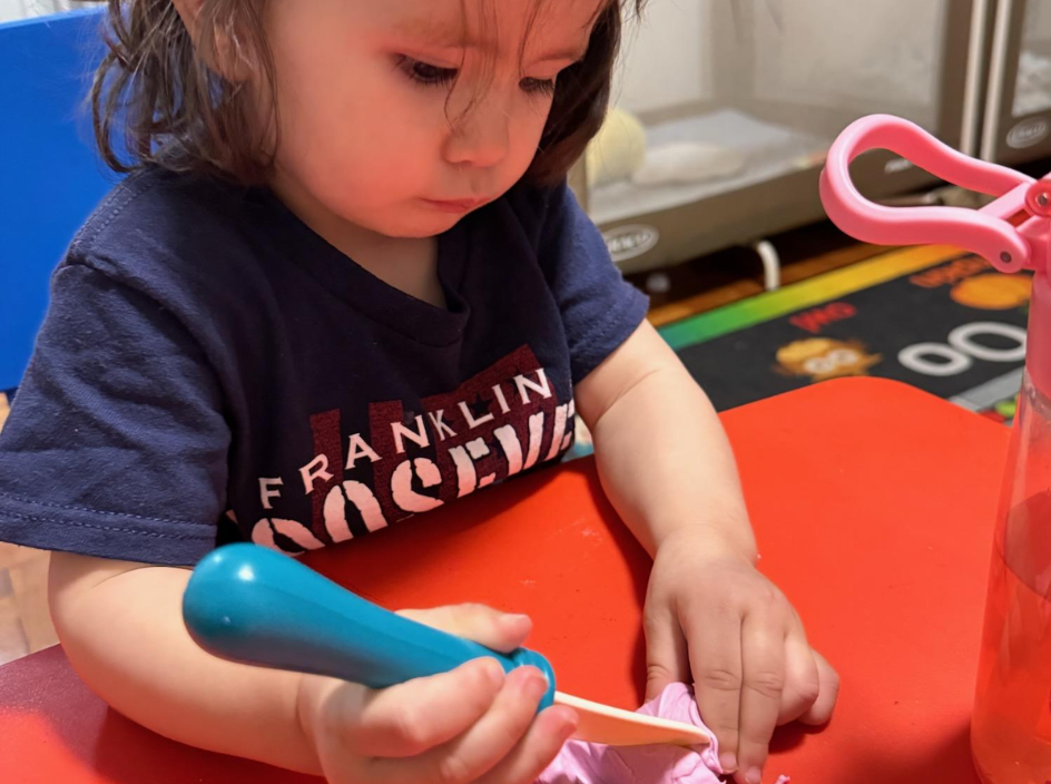 A preschooler shaping pink play dough with a craft stick at a table
