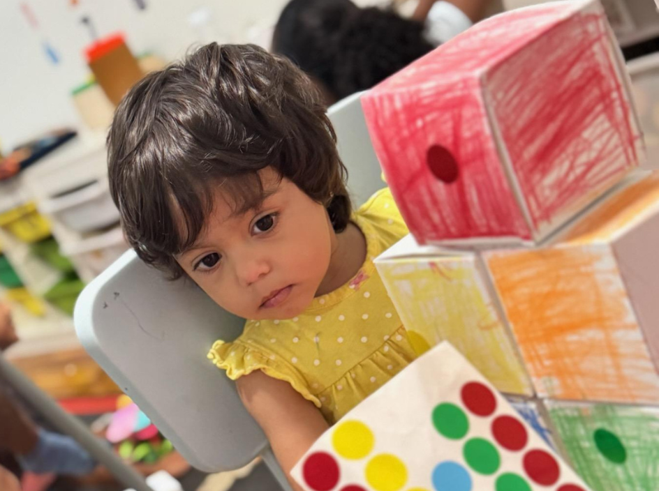 A toddler in a yellow dress playing with colorful hand-decorated cardboard boxes